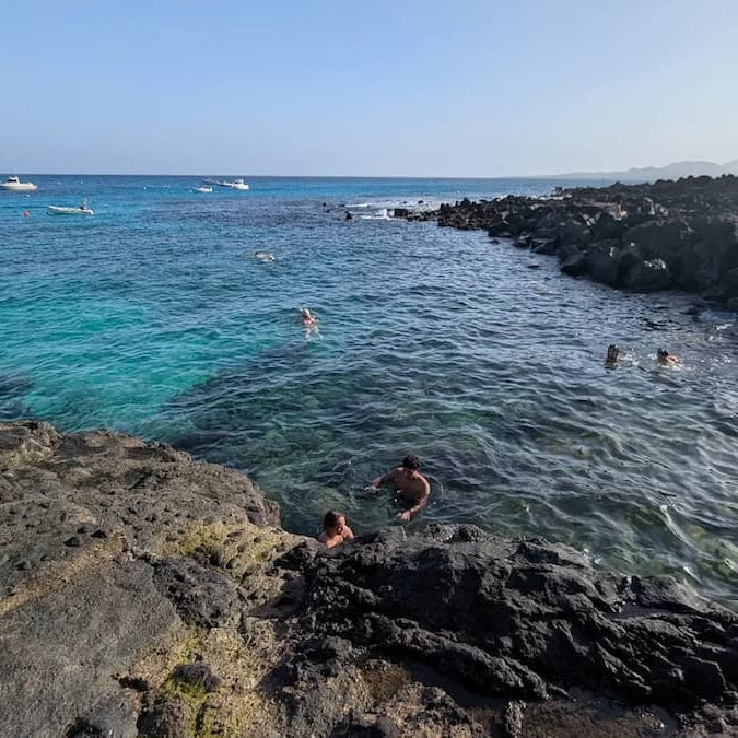 People swimming and playing in a rocky coastal area with clear blue water and a clear sky.