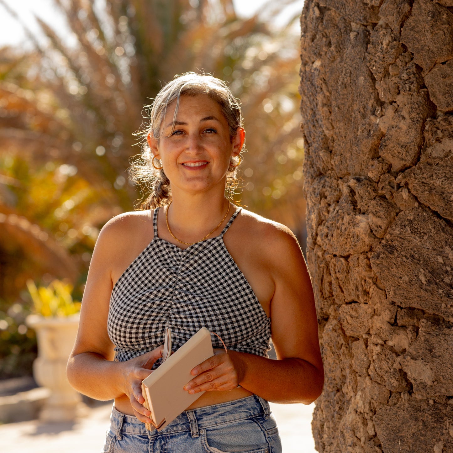 Lucia holding a sketchbook outdoors near a stone wall with palm trees in the background
